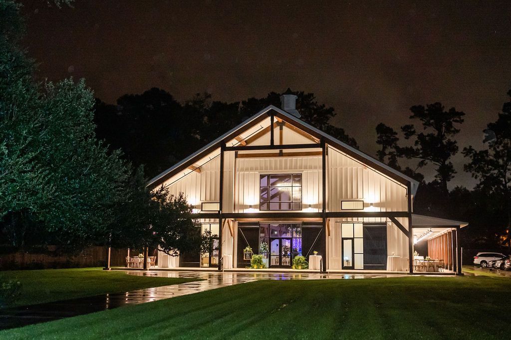 A large illuminated barn at night; wooden structure with lights, glass windows, and green lawn.