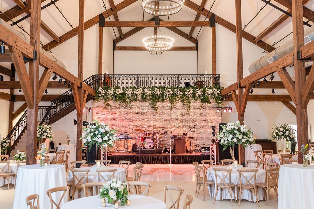 Reception hall with wooden beams, tables with floral centerpieces, stage with band, chandelier.