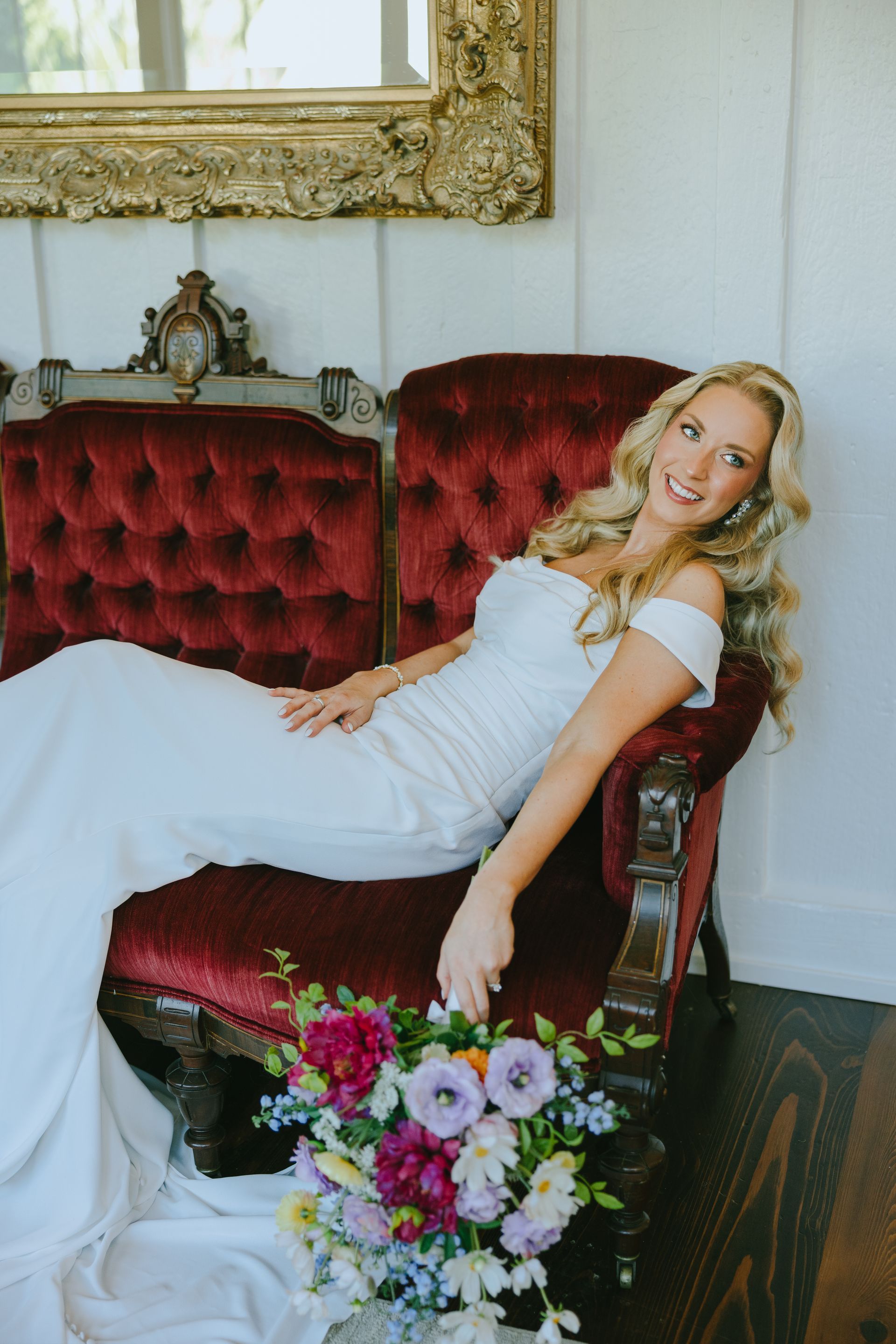 Woman in white dress smiles, reclines on red velvet chair with bouquet; ornate mirror in background.