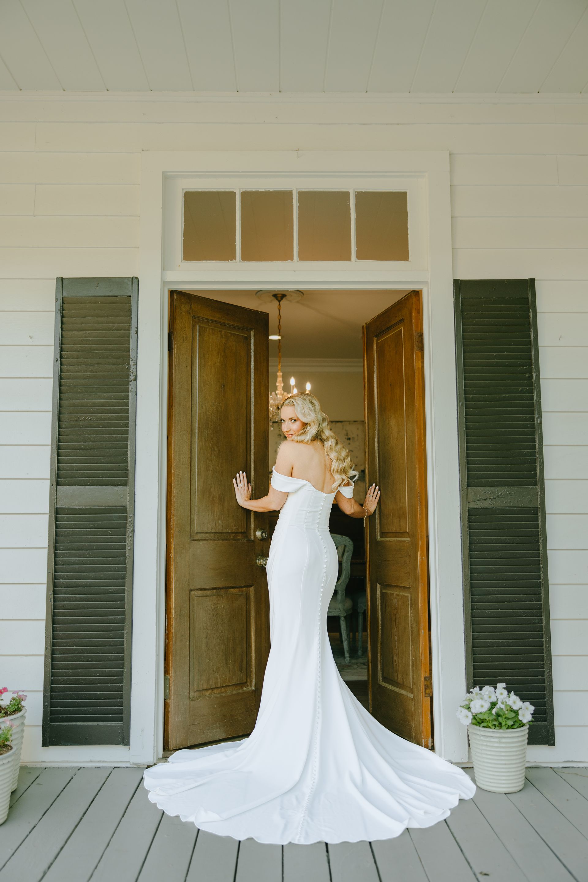 Bride in a white gown stands in a doorway, back to the viewer, holding the door open on a porch.
