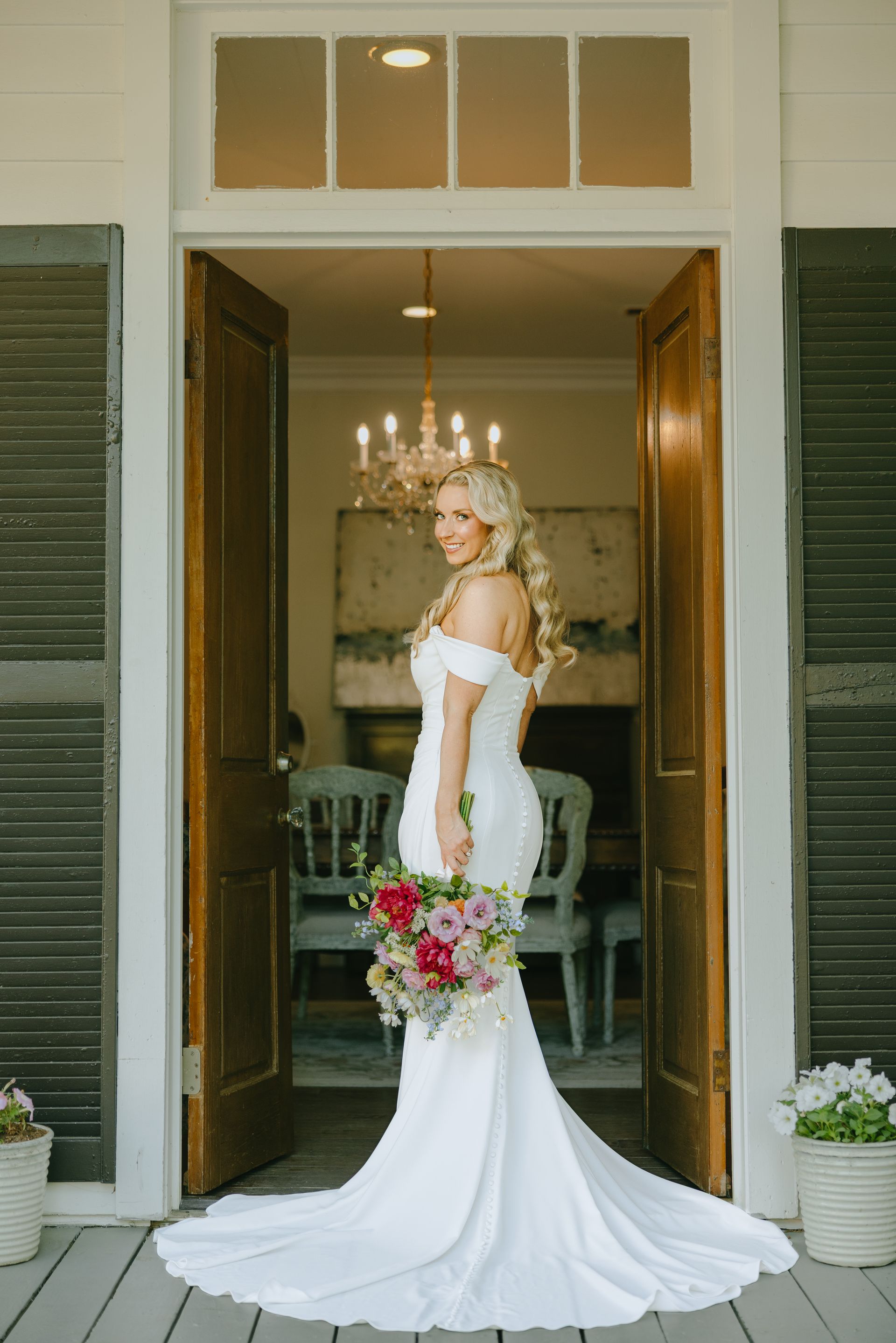 Bride in a white off-the-shoulder gown, holding a bouquet, standing in a doorway.
