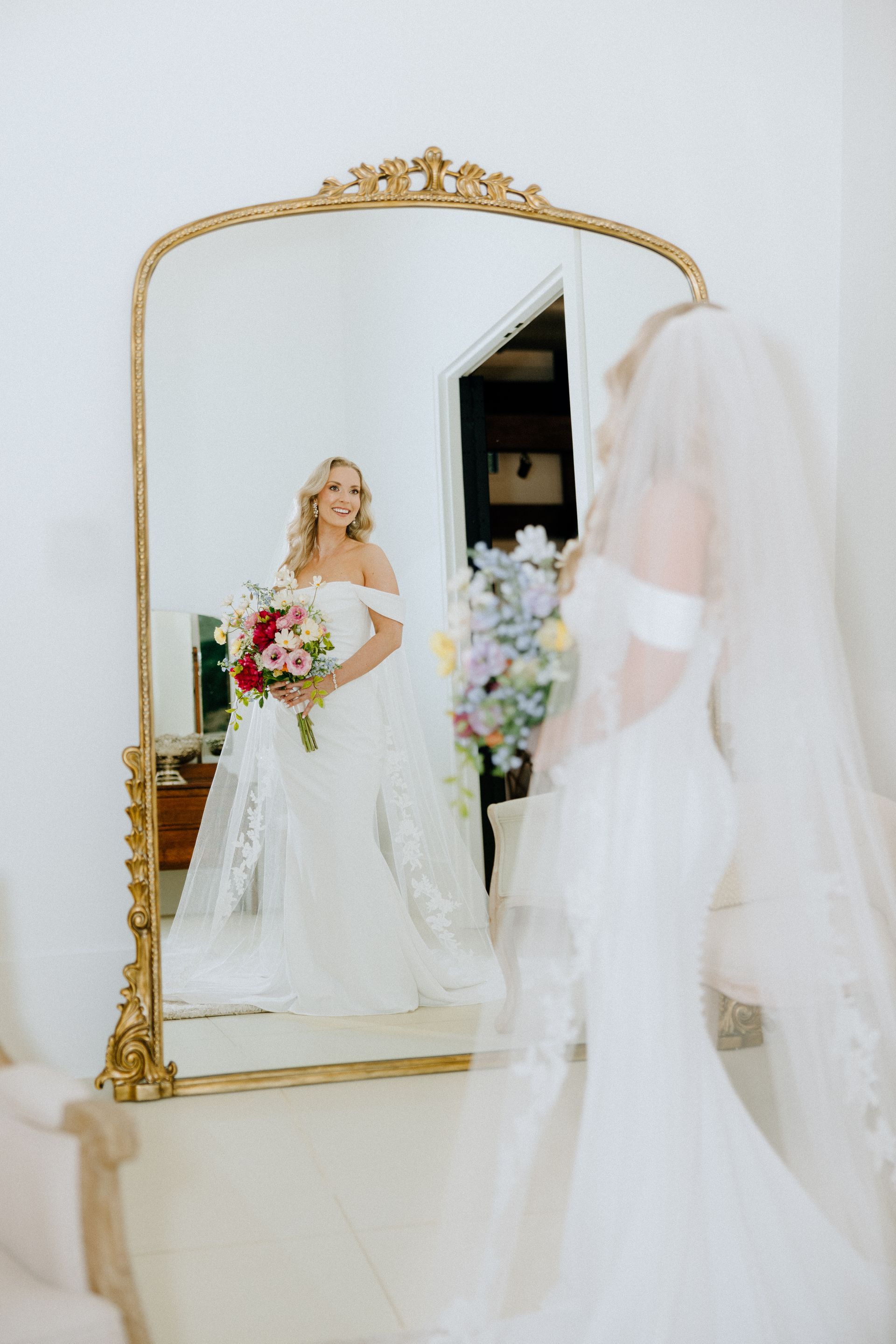 Bride in wedding dress, looking in gilded mirror, holding bouquet of flowers.