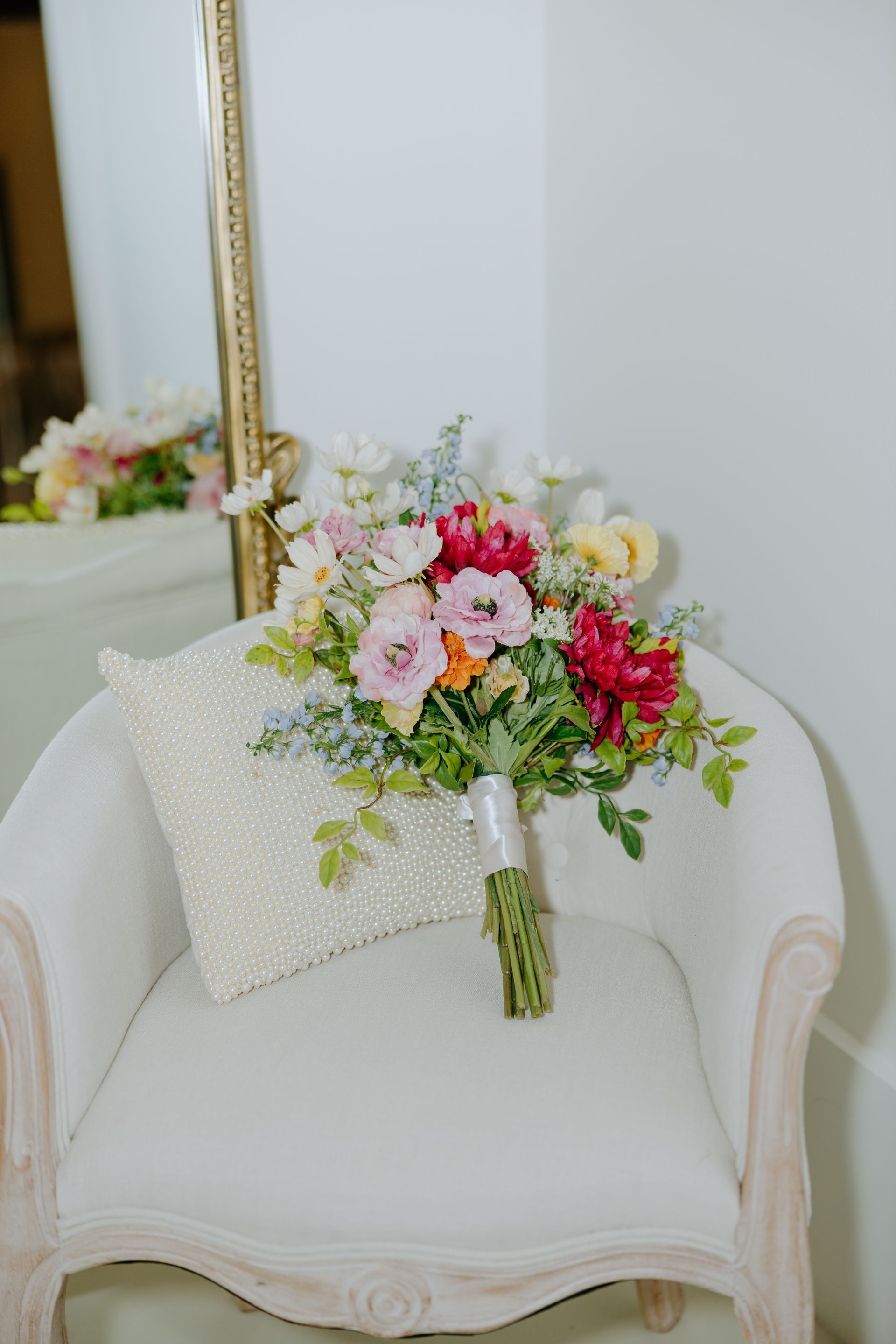 A colorful floral bouquet rests on a white chair next to a decorative pillow. A gold-framed mirror is visible.