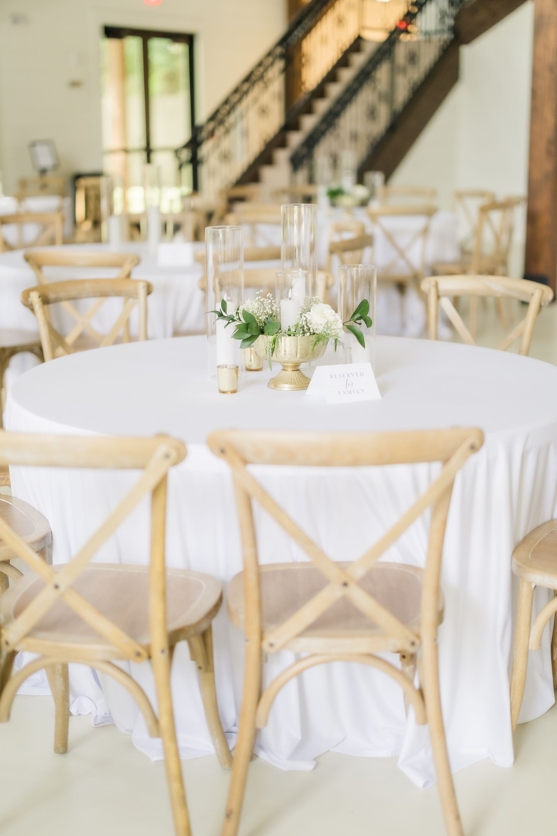 Round table set for a wedding, with white linens, floral centerpiece, and wooden cross-back chairs.