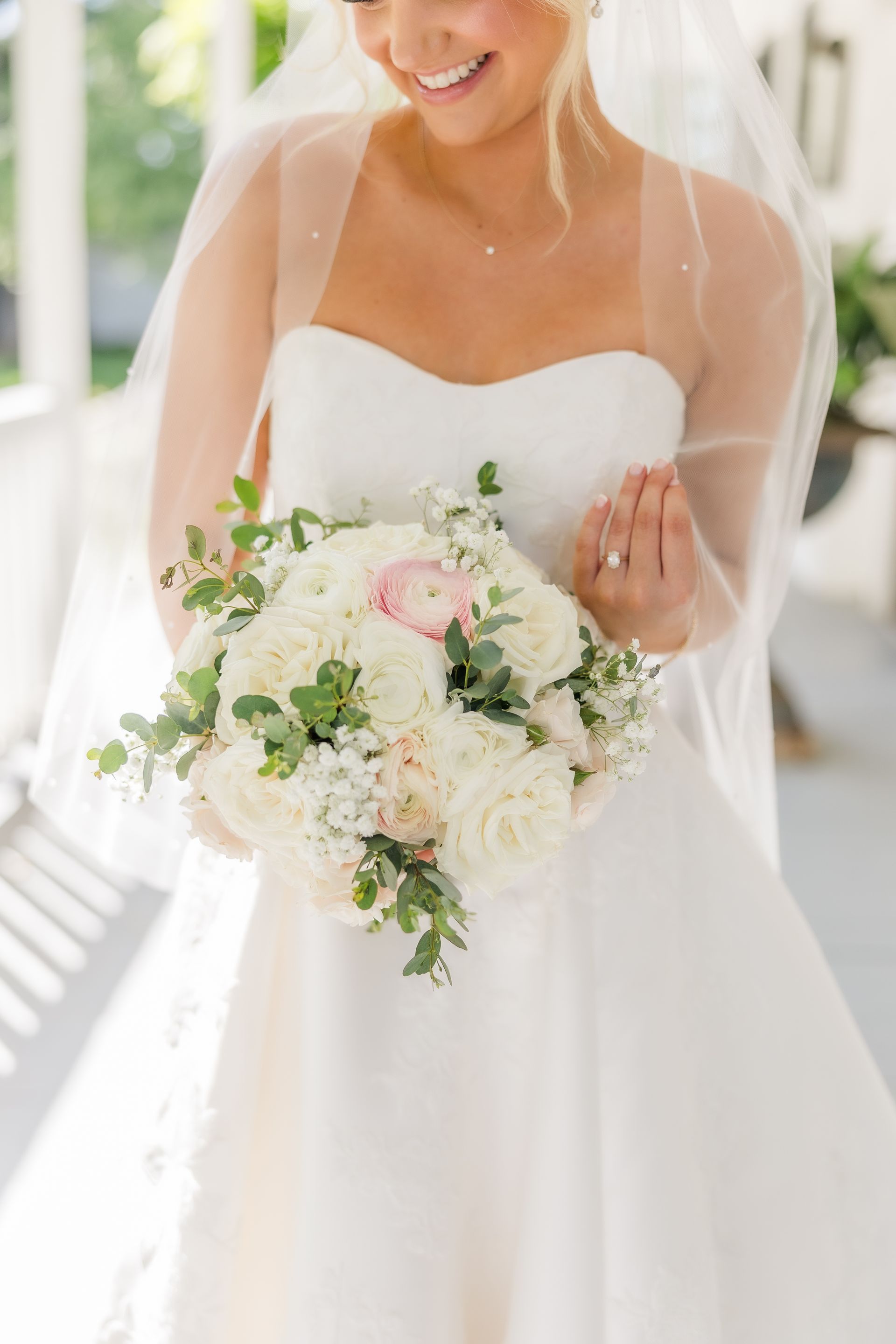 Bride holding a bouquet, wearing a strapless white dress, smiling, with a sheer veil.