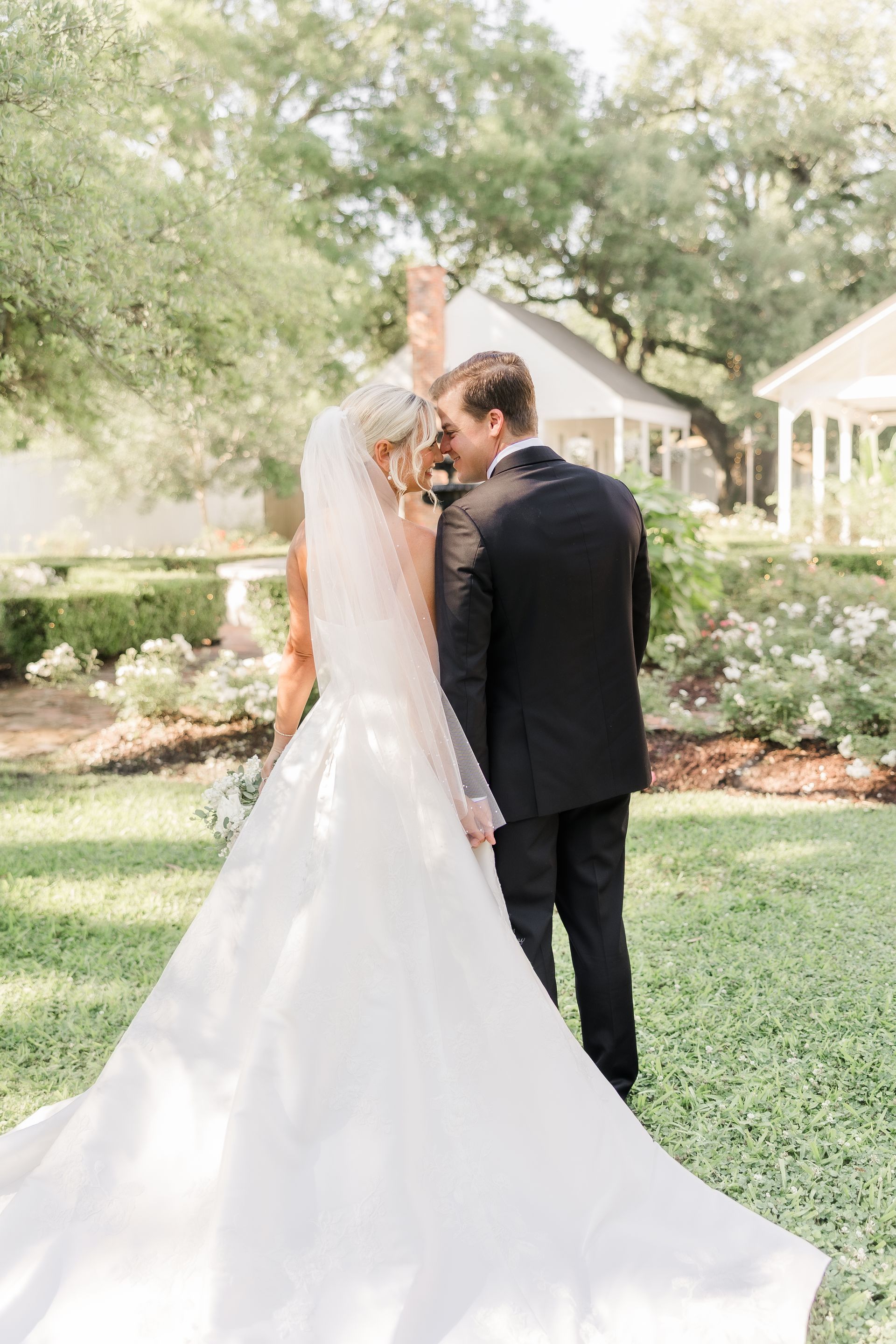 Bride and groom embrace in a garden. The bride wears a white gown and veil; the groom wears a black suit.
