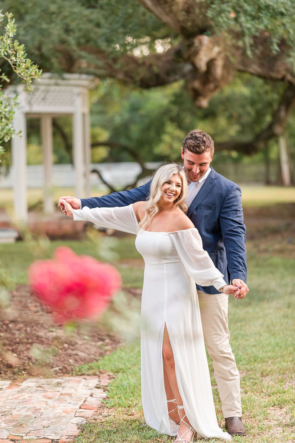 Couple dancing outdoors near a white gazebo. Woman in white dress smiles, man in navy blazer.