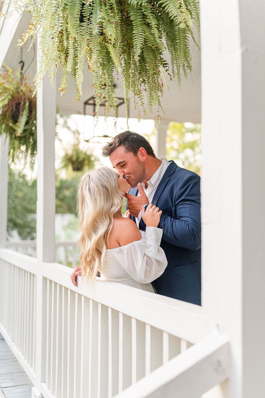 Couple kissing on a porch, white railing, hanging plants, man in blue blazer, woman in white dress.