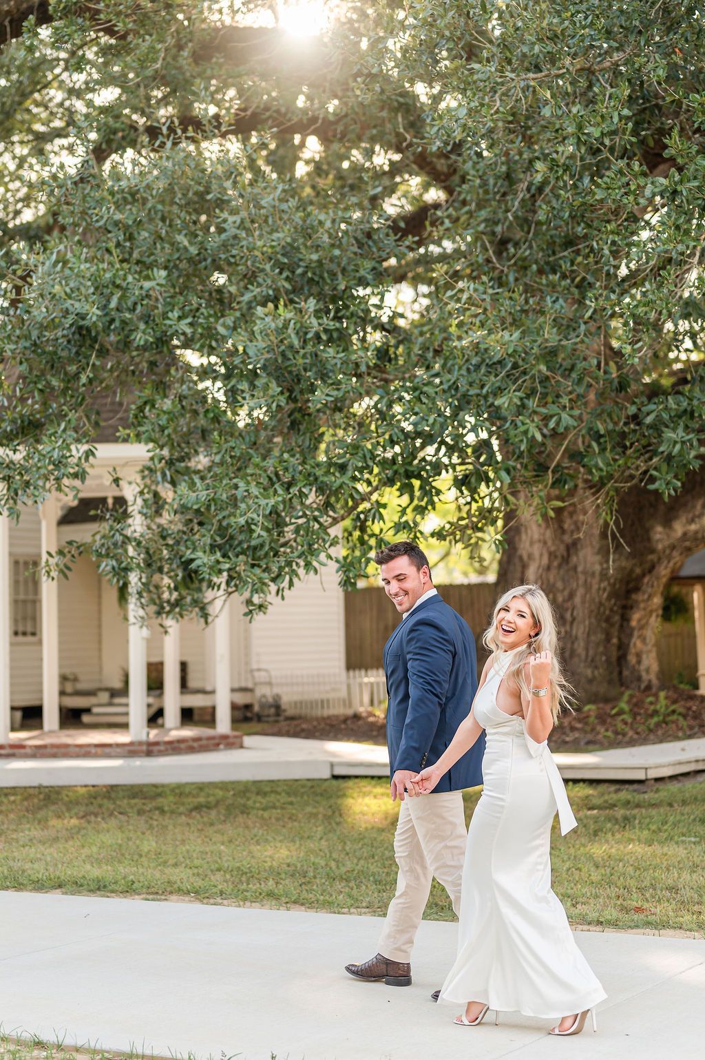 Couple walking hand-in-hand toward camera in front of a white building and large tree; sunny outdoor setting.