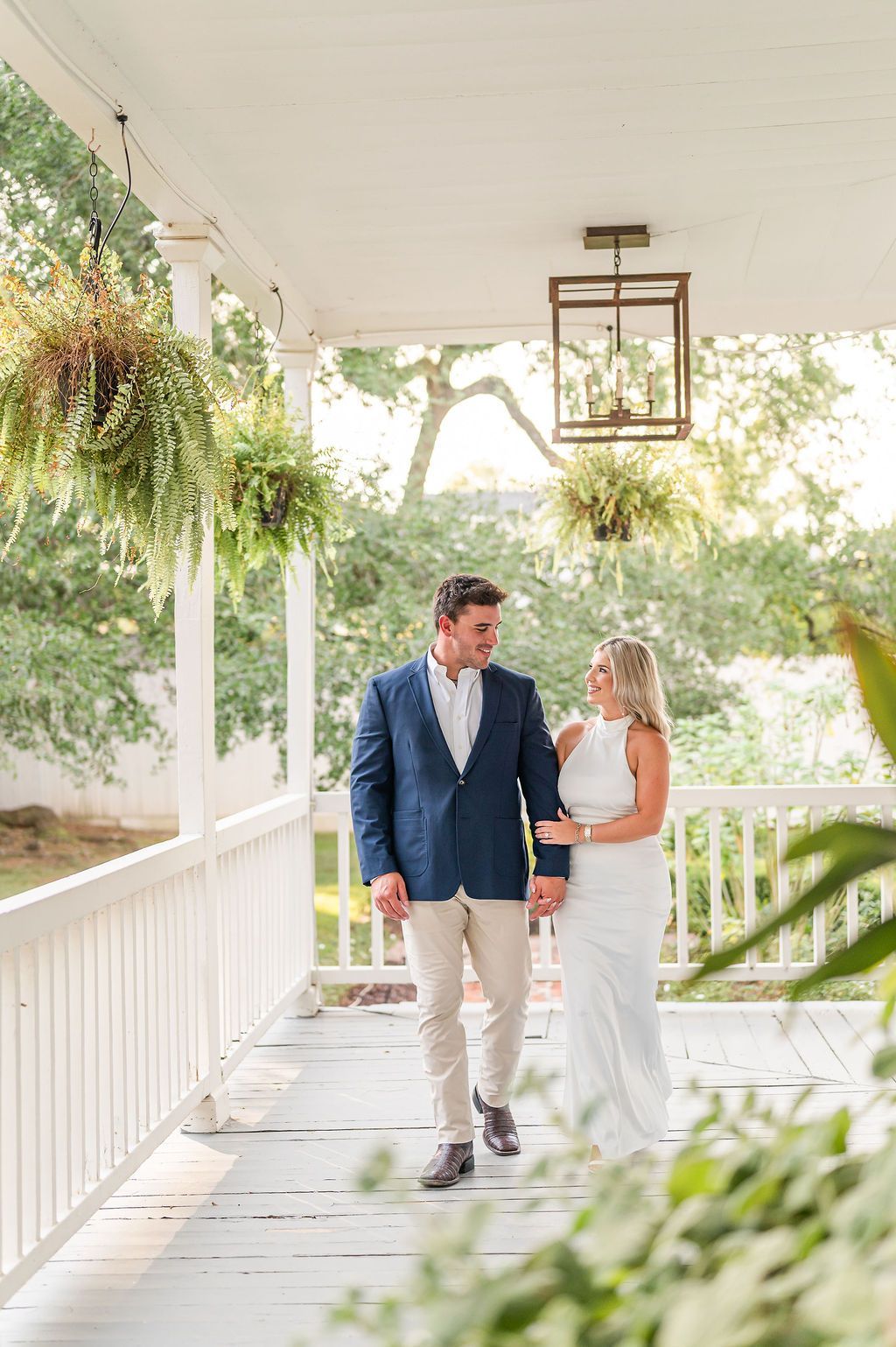 Couple holding hands, walking on a porch. The man wears a blazer and khakis, the woman a white dress.