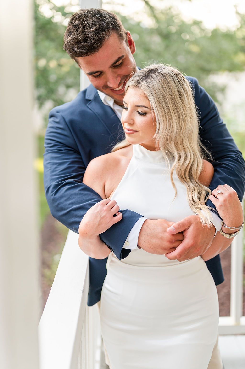 Man in blue blazer hugs woman in white dress on a balcony, both smiling.