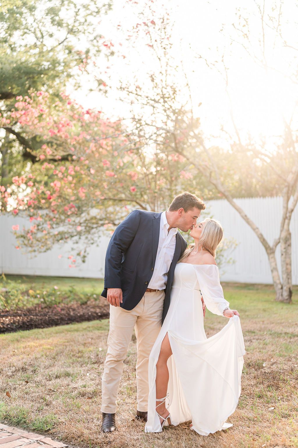 Couple kissing outdoors, woman in white dress, man in blazer, sunny garden setting.