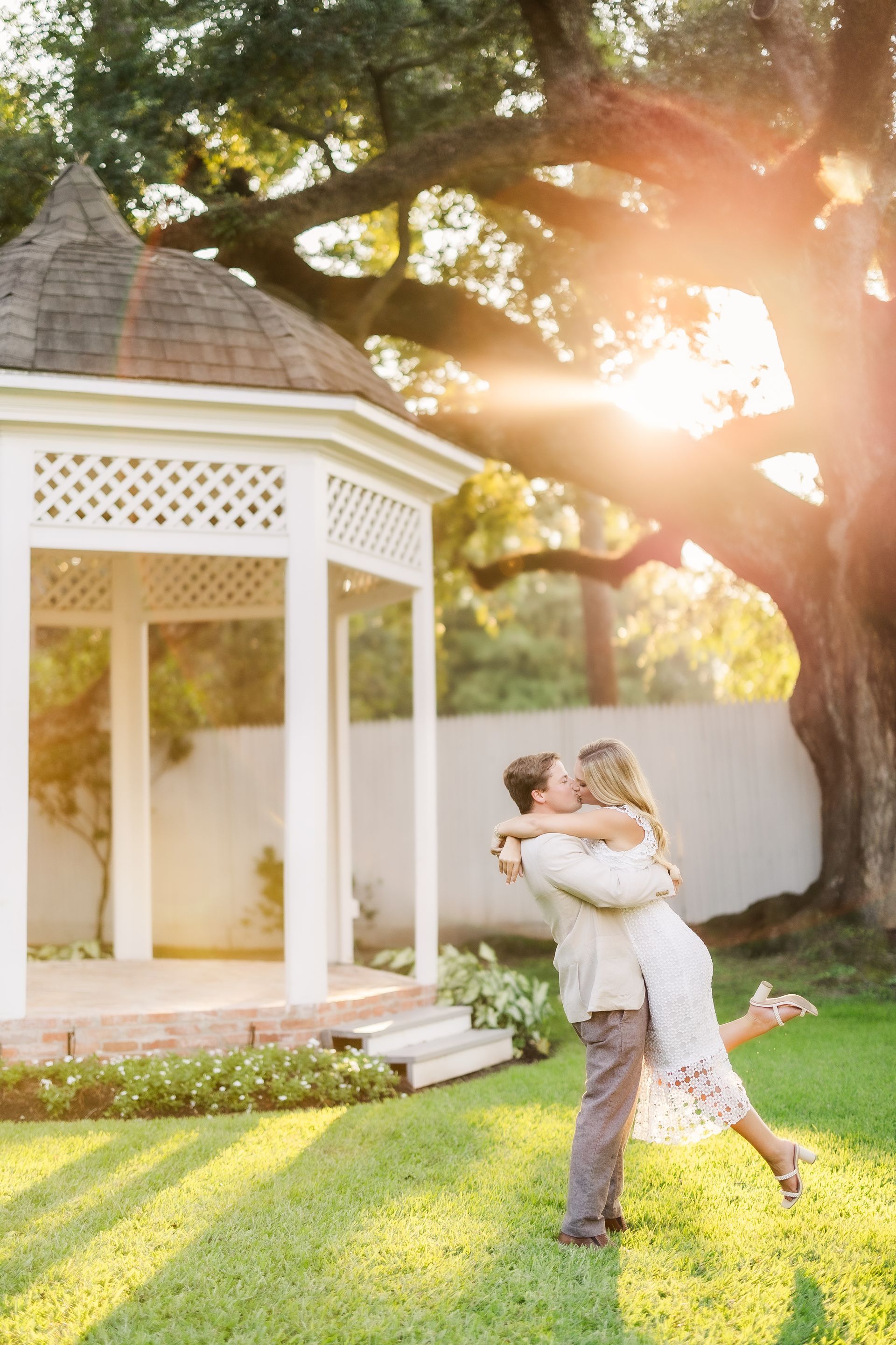 Man lifting woman, kissing, near gazebo in sunny garden.