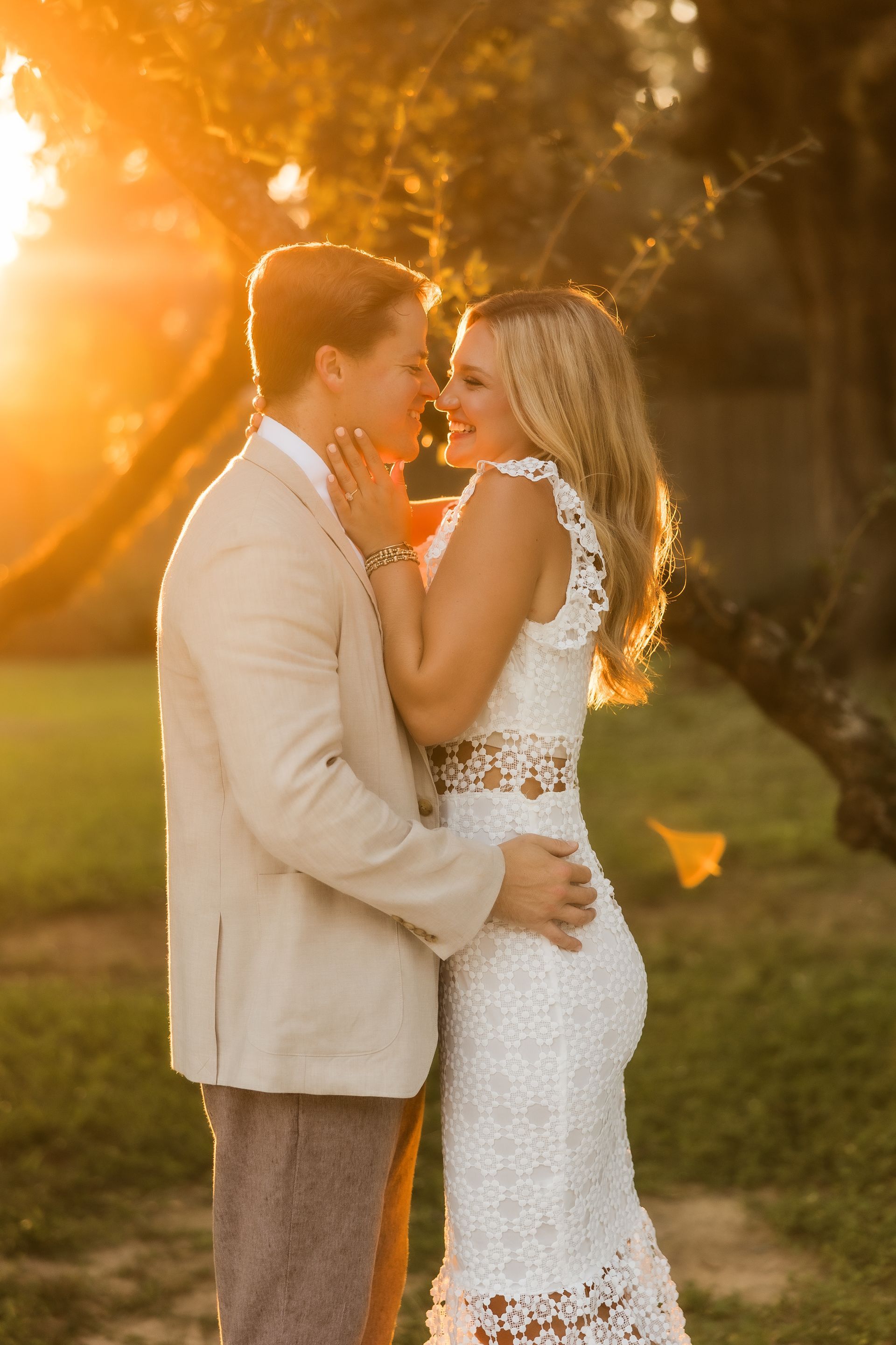 Couple embraces, bathed in golden sunlight. Woman in white lace dress, man in blazer, outdoors.