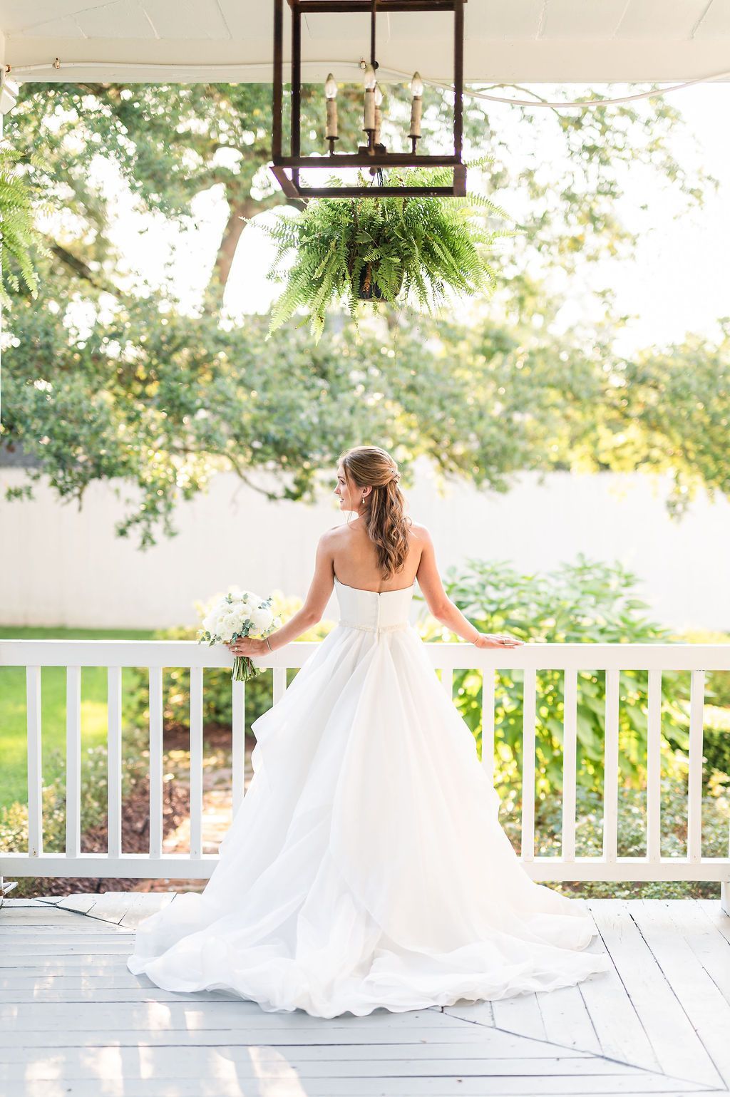 Bride in white wedding dress on a porch, holding flowers, gazing at a sunlit garden.