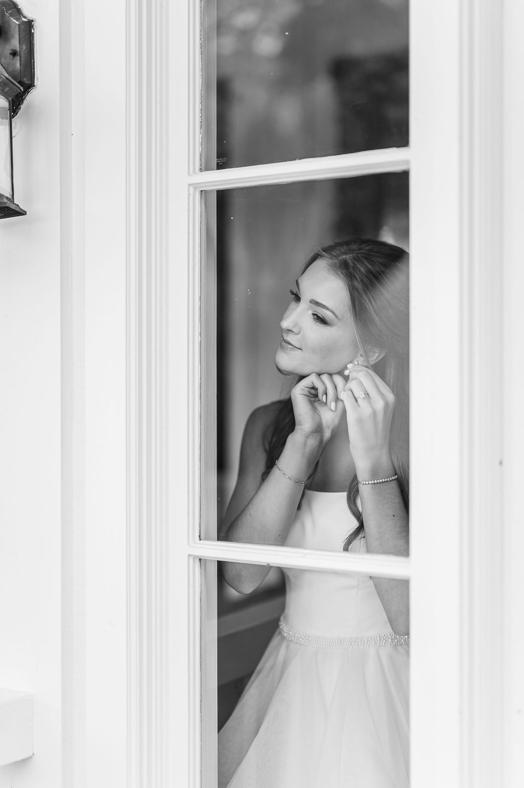Woman in a wedding dress, looking in a window, putting on earrings.