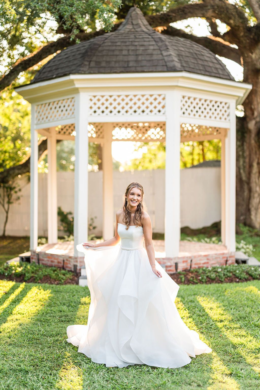 Woman in white dress smiles, stands near a gazebo in a sunny garden.