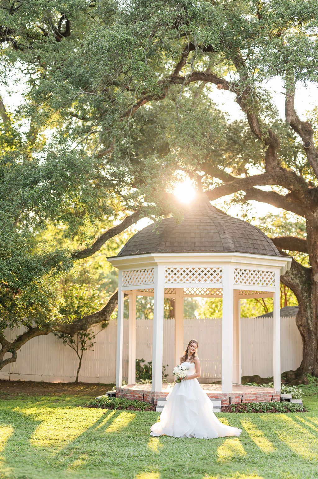 Bride in a white gown holding a bouquet stands in a gazebo in a sunny garden.