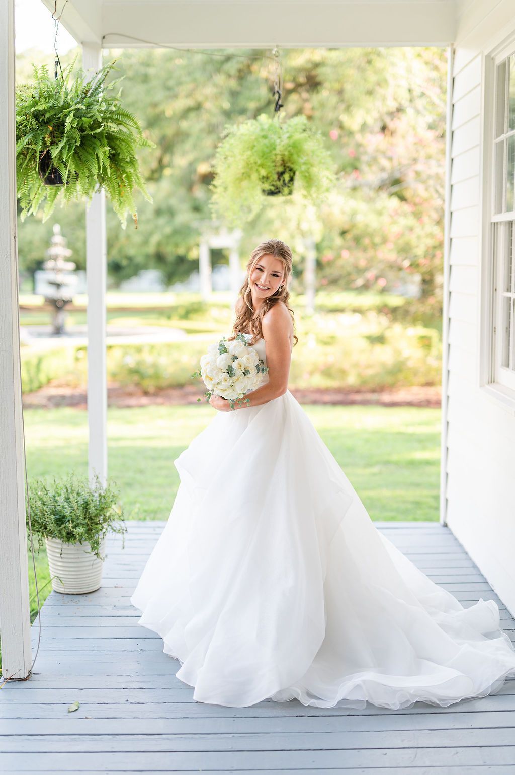 Bride in a white gown holding a bouquet smiles on a porch with ferns.