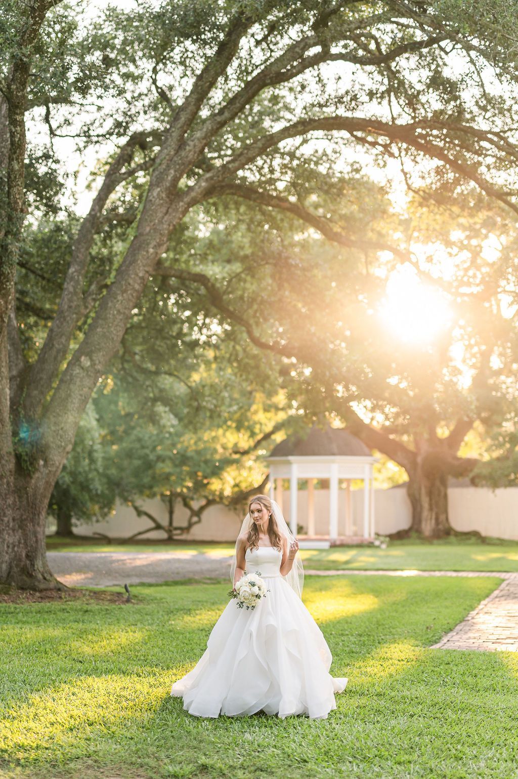 Bride in white dress holding flowers, standing in sunlit garden near a gazebo.