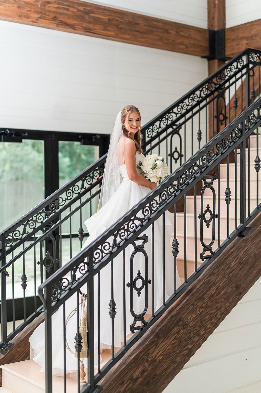 Bride in white dress holding flowers, on a staircase with black railing and wooden accents.