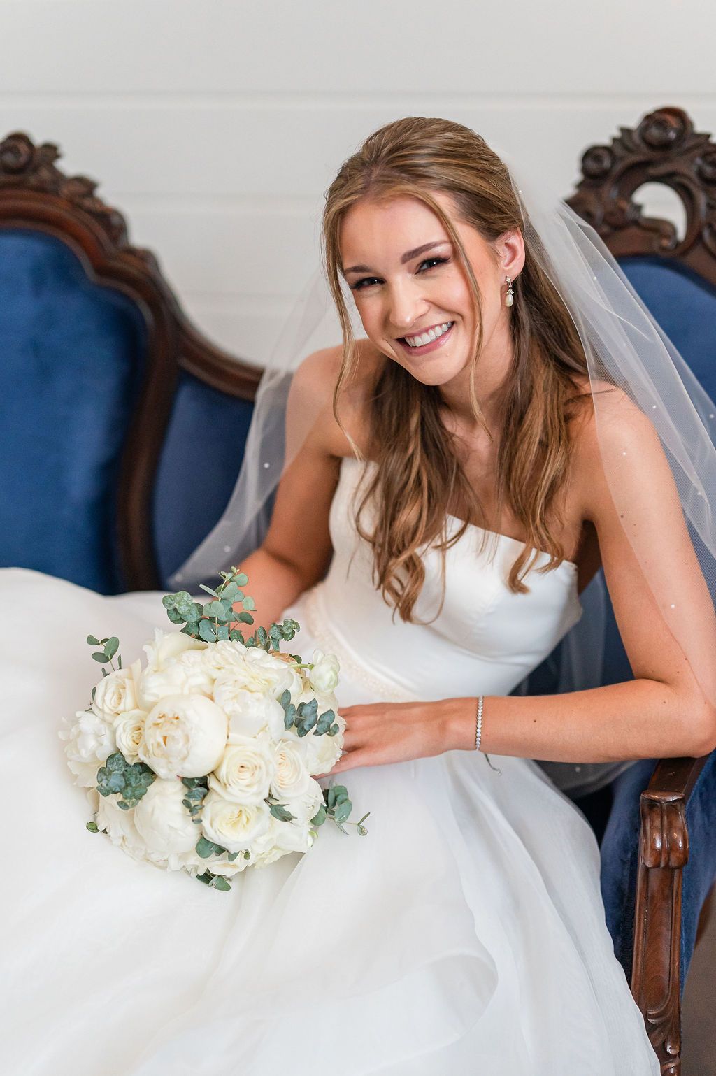 Bride in strapless white gown, holding bouquet, smiles while seated on blue ornate chair.