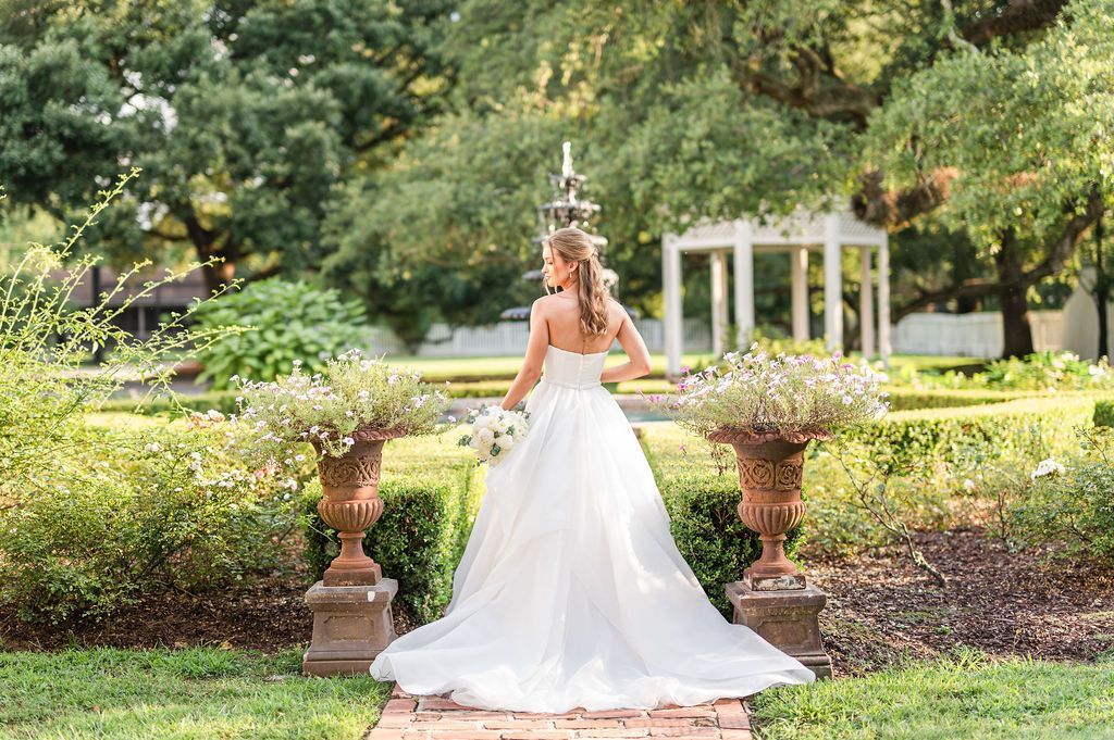 Bride in a white gown stands in a garden, holding flowers, facing away from the camera.
