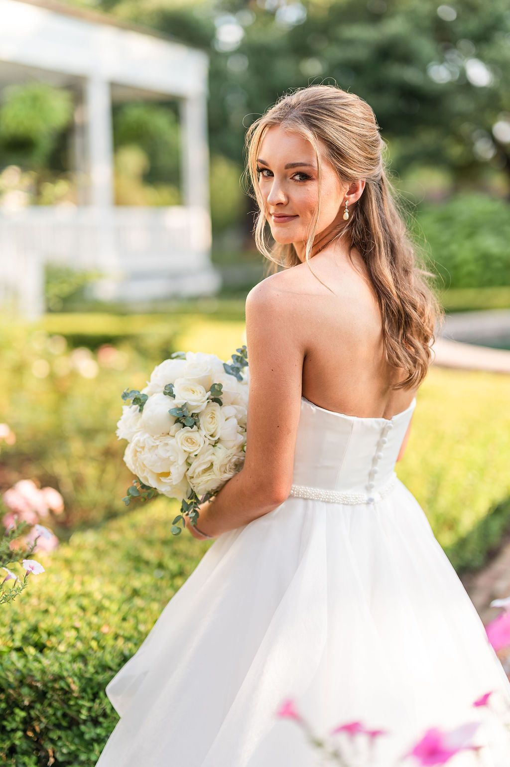 Bride in a white strapless gown holds a bouquet, looking over her shoulder, in a garden.