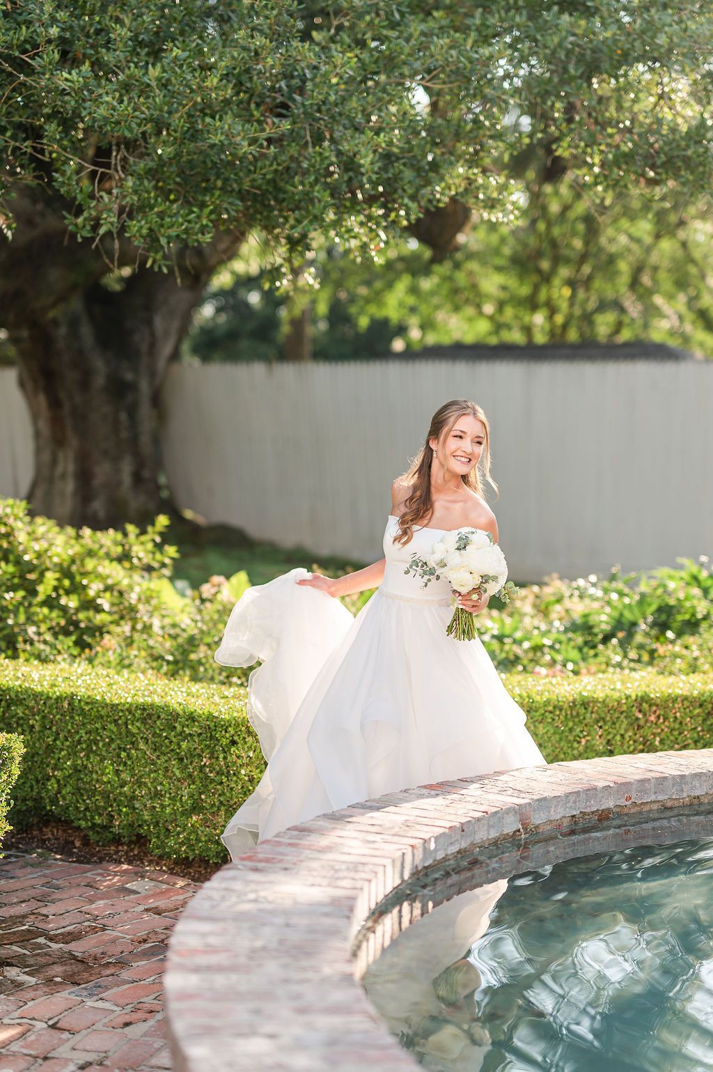 Bride in a white gown holds bouquet, stands by a fountain in a garden.