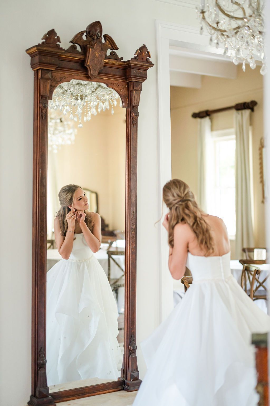 Woman in white gown putting on earrings, reflected in ornate wooden mirror.