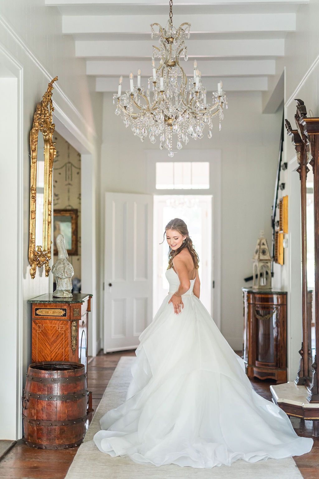 Bride in a white gown stands in hallway, looking back. Chandelier hangs overhead, antique furniture and mirror.