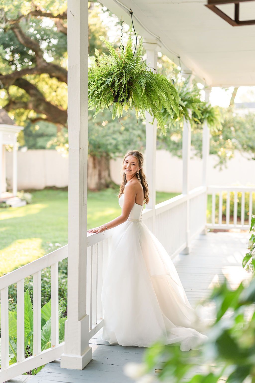 Bride in strapless gown poses on a white porch, greenery overhead.