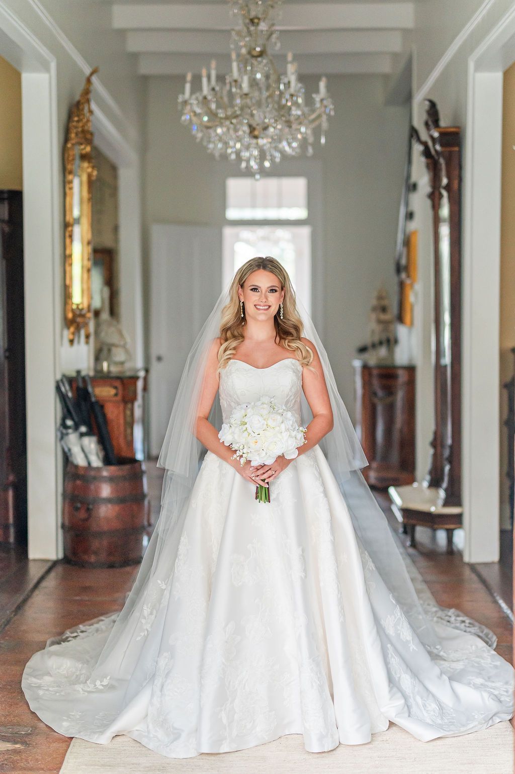 Bride in white strapless gown holds flowers, standing in a hallway under a crystal chandelier.