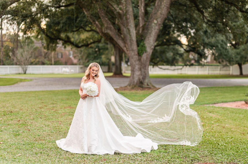 Bride in a white gown and veil holds a bouquet, standing in a grassy yard under a large tree.