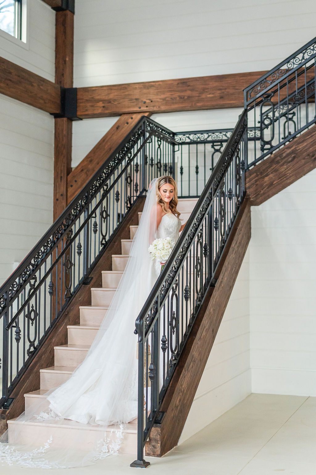 Bride in a white gown and veil stands on a staircase, holding flowers. The interior has wood beams and wrought iron.