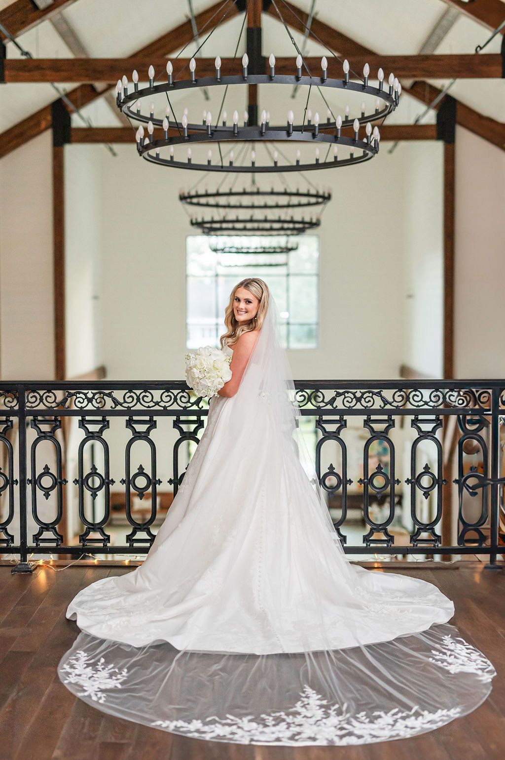 Bride in a white gown holding bouquet, smiling. Standing on a balcony with chandelier lights overhead.