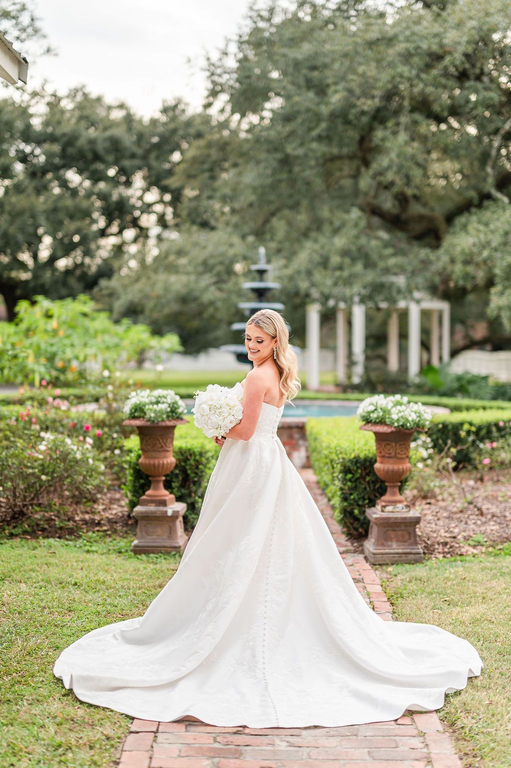 Bride in white gown holding bouquet, posing in a garden with fountain and brick walkway.
