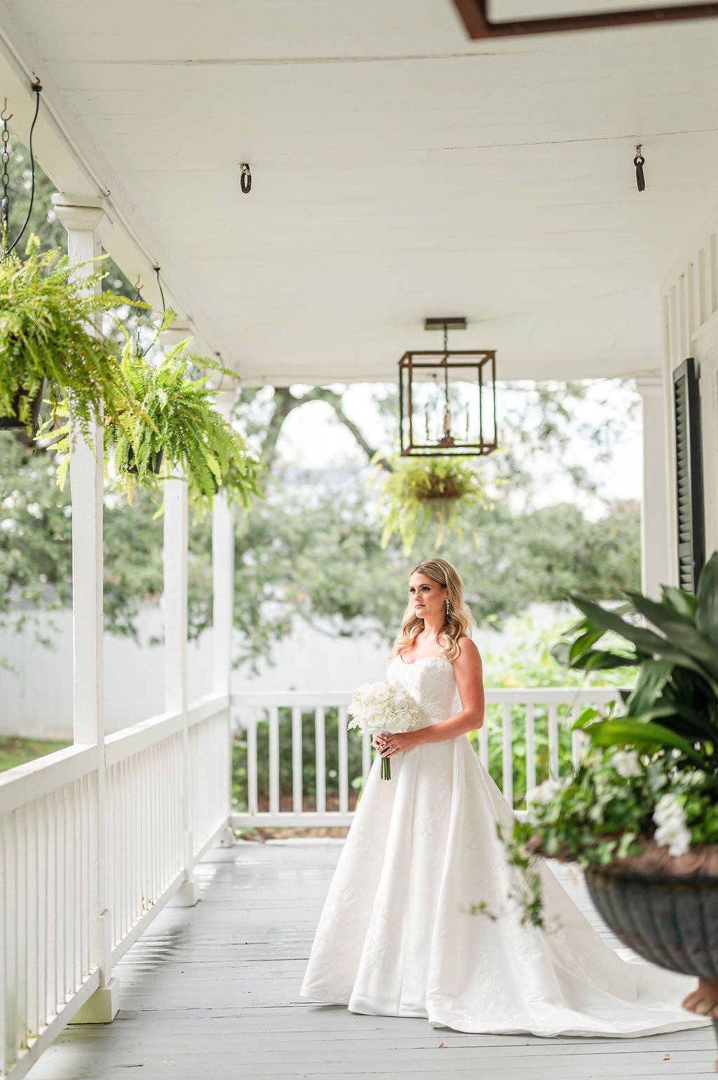 Bride in a white gown holding flowers, standing on a porch with ferns and a chandelier.