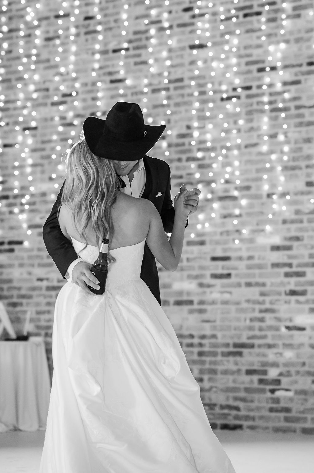 Bride and groom dance at reception; groom wears a cowboy hat, and fairy lights hang behind a brick wall.