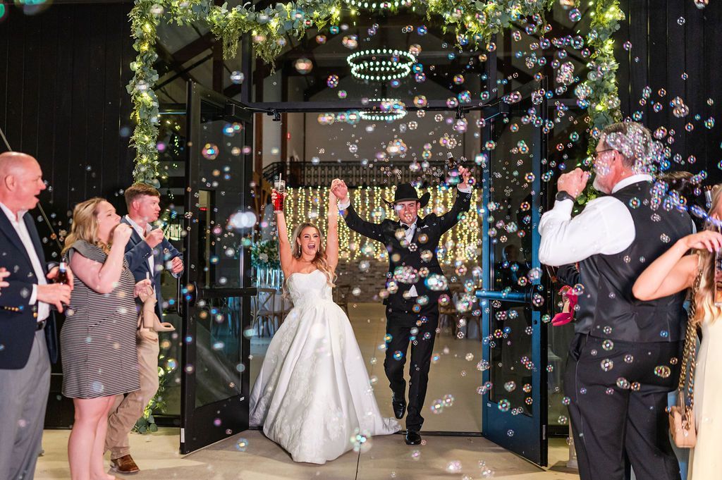Newlyweds celebrate their exit, raising arms, surrounded by bubbles. Guests cheer, decorated doorway.