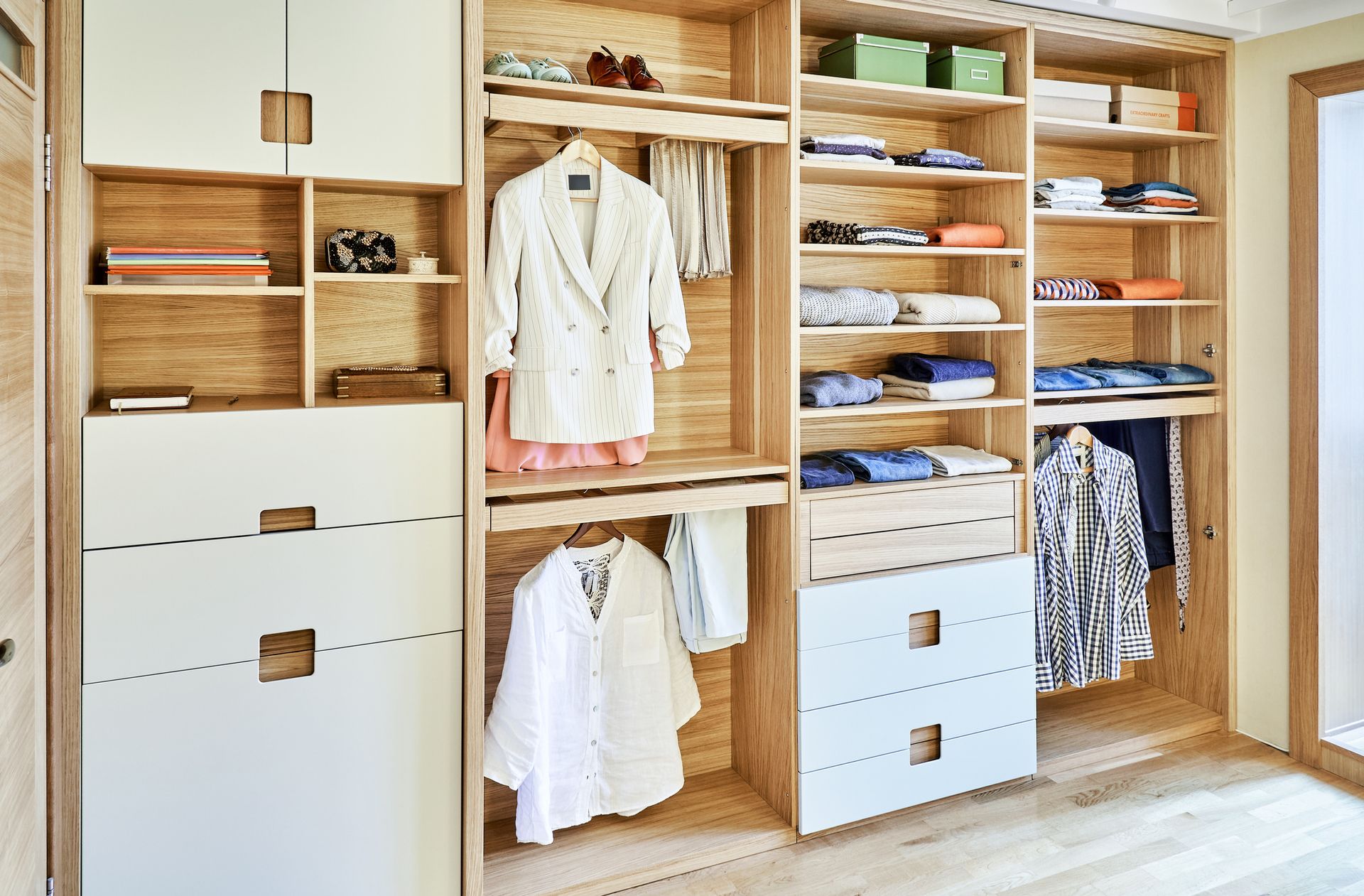 Wooden walk-in closet with white and natural wood shelving, drawers, and hanging rods, displaying clothing and accessories.
