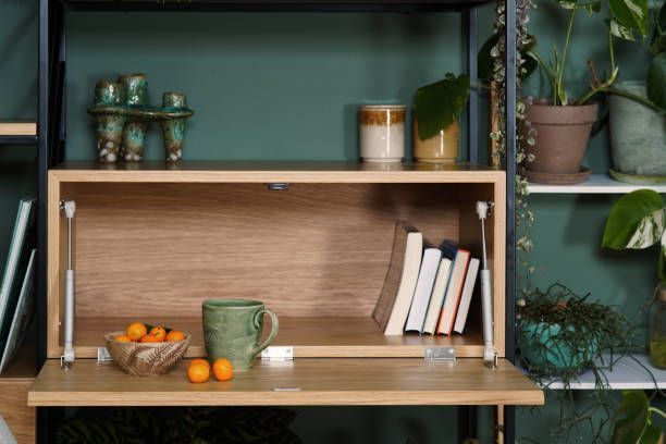 Desk with open wooden drop-down desk area, books, mug, oranges, with a shelf above holding decor and plants.