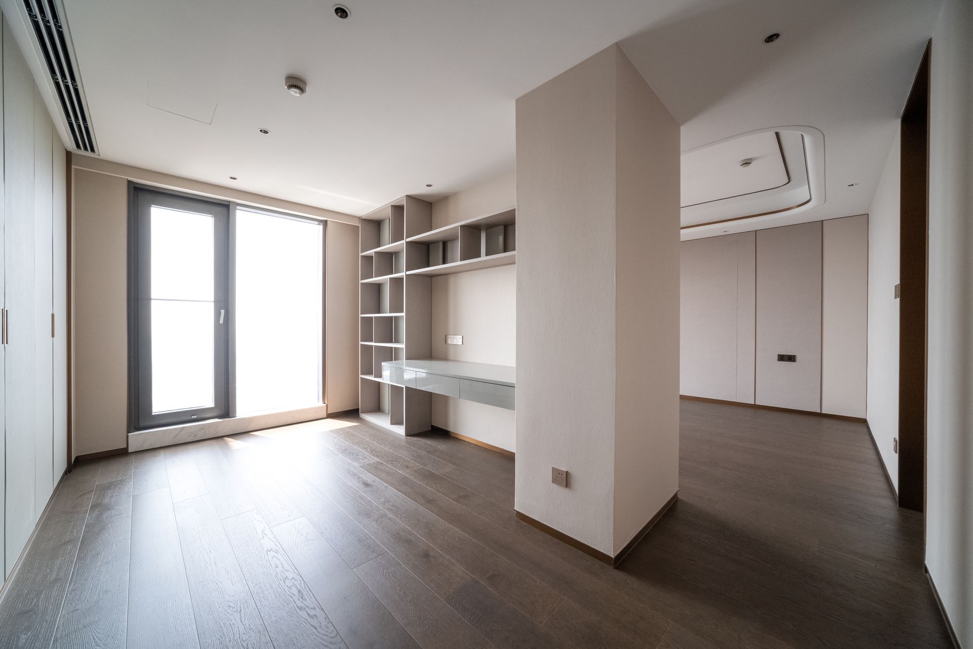 Empty modern room with wood floor, built-in shelves, large window, and a light-colored desk.