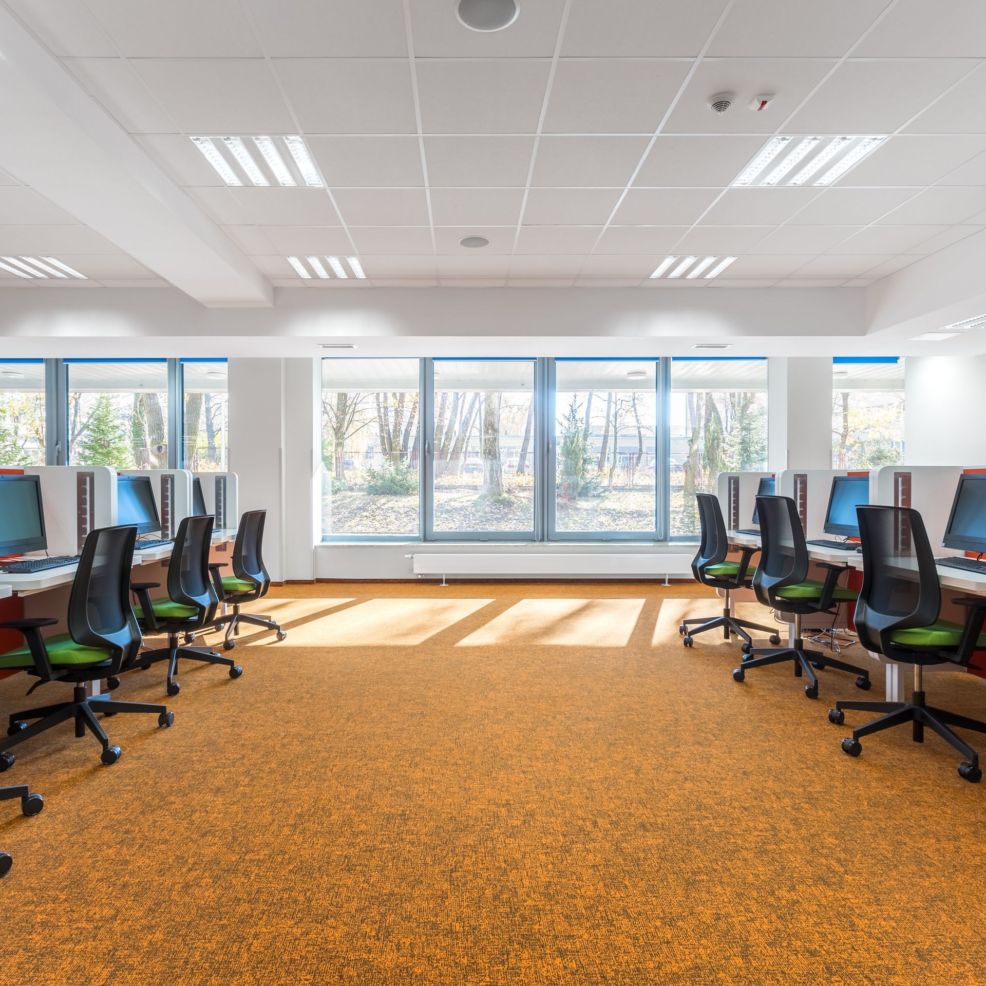 Computer lab with orange carpet, computers, and ergonomic chairs; windows in background.
