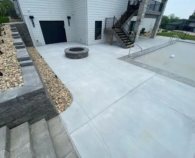 Concrete patio with fire pit, stairs, and pool adjacent to a white house with black garage door and exterior stairs.