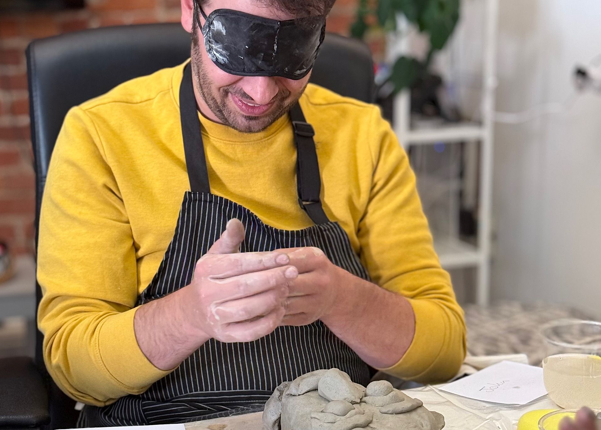 A blindfolded man is making a clay pot at a table.