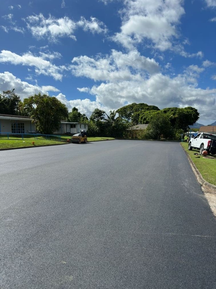 Newly paved asphalt road on a sunny day, flanked by grass and houses under a partly cloudy sky.