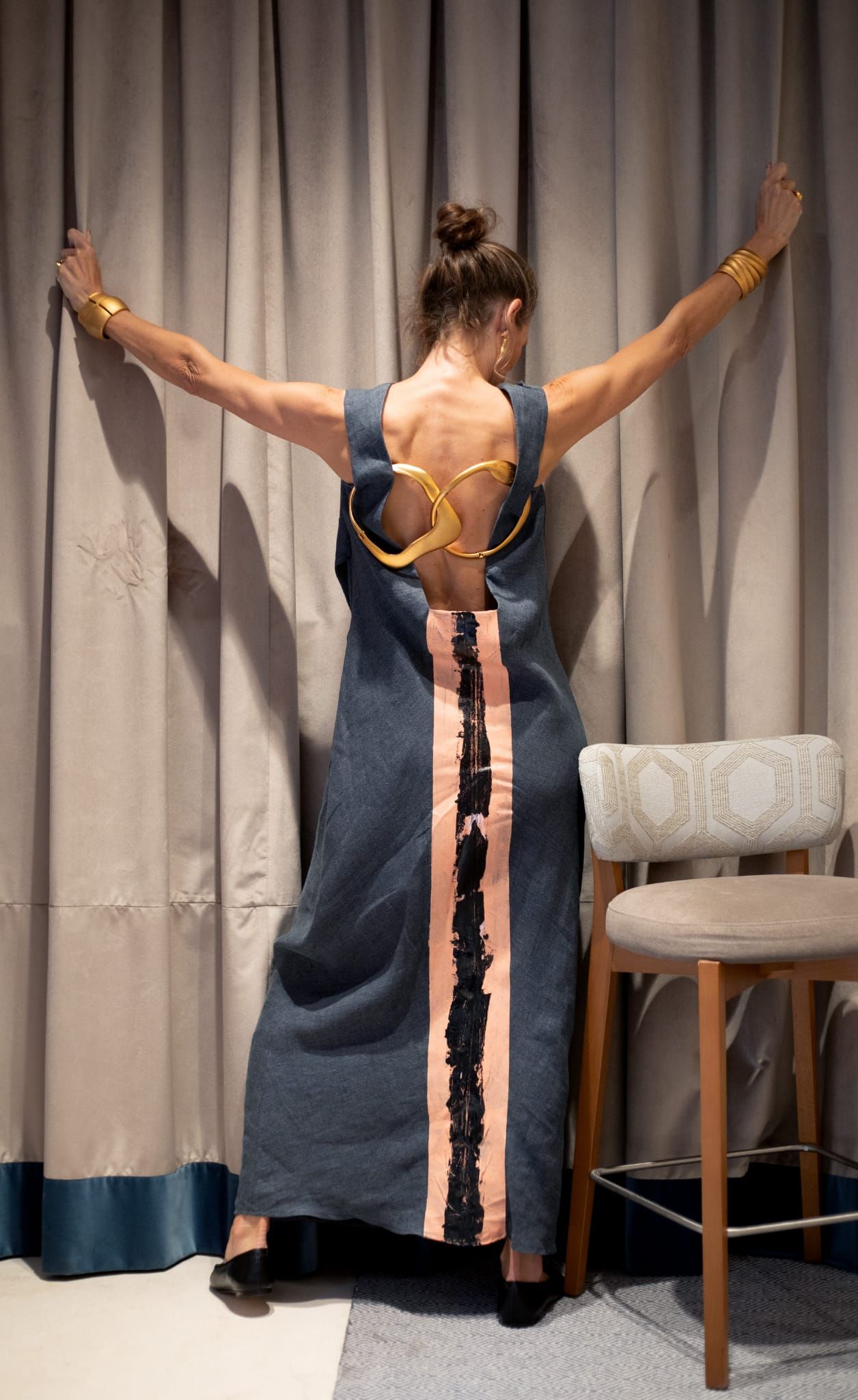 Mujer con vestido sin espalda y joyas de oro, con los brazos extendidos, apoyada en unas cortinas.