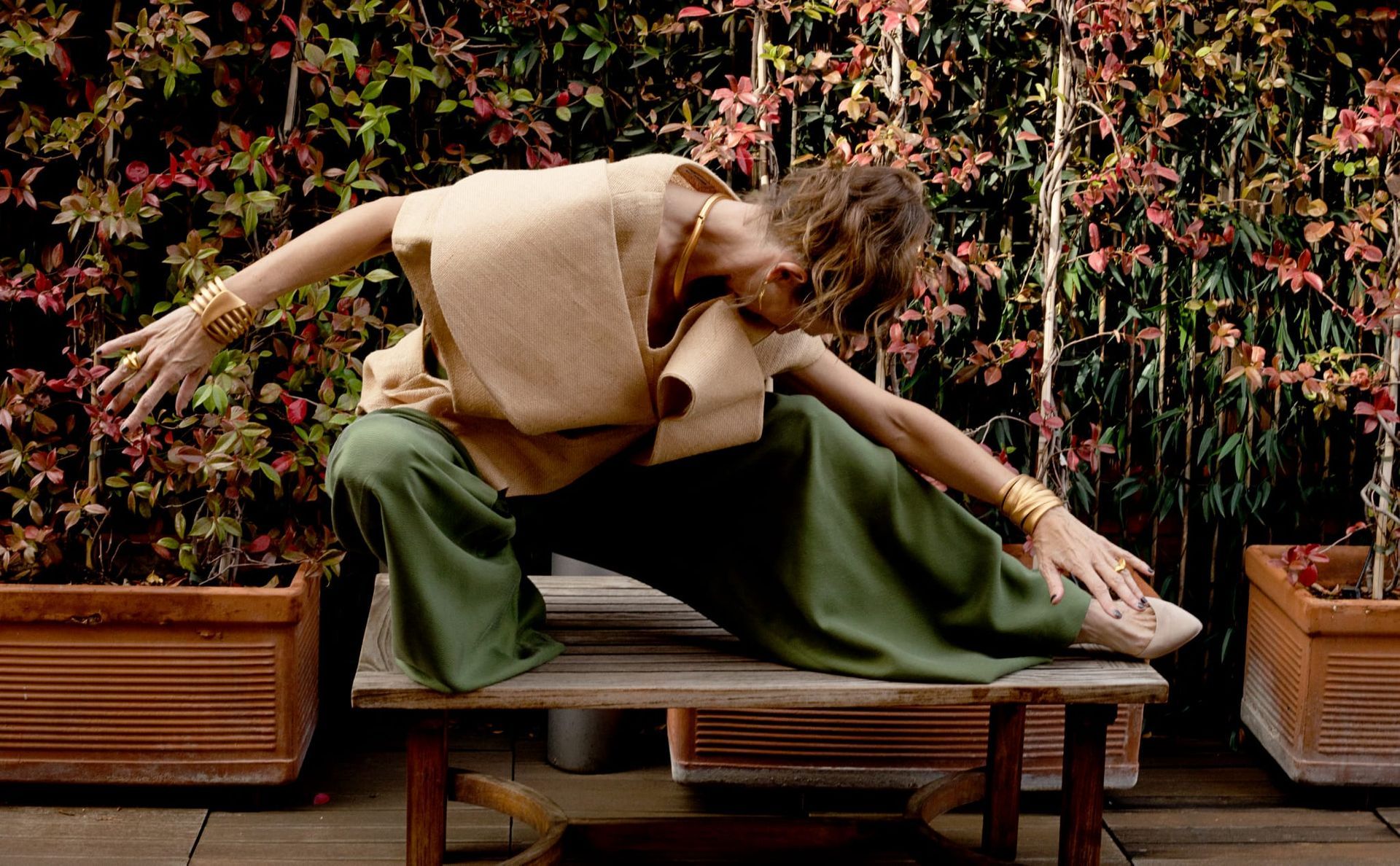 Una mujer con pantalones verdes y blusa color canela se estira en un banco de madera, con los brazos extendidos, al aire libre cerca de la vegetación.