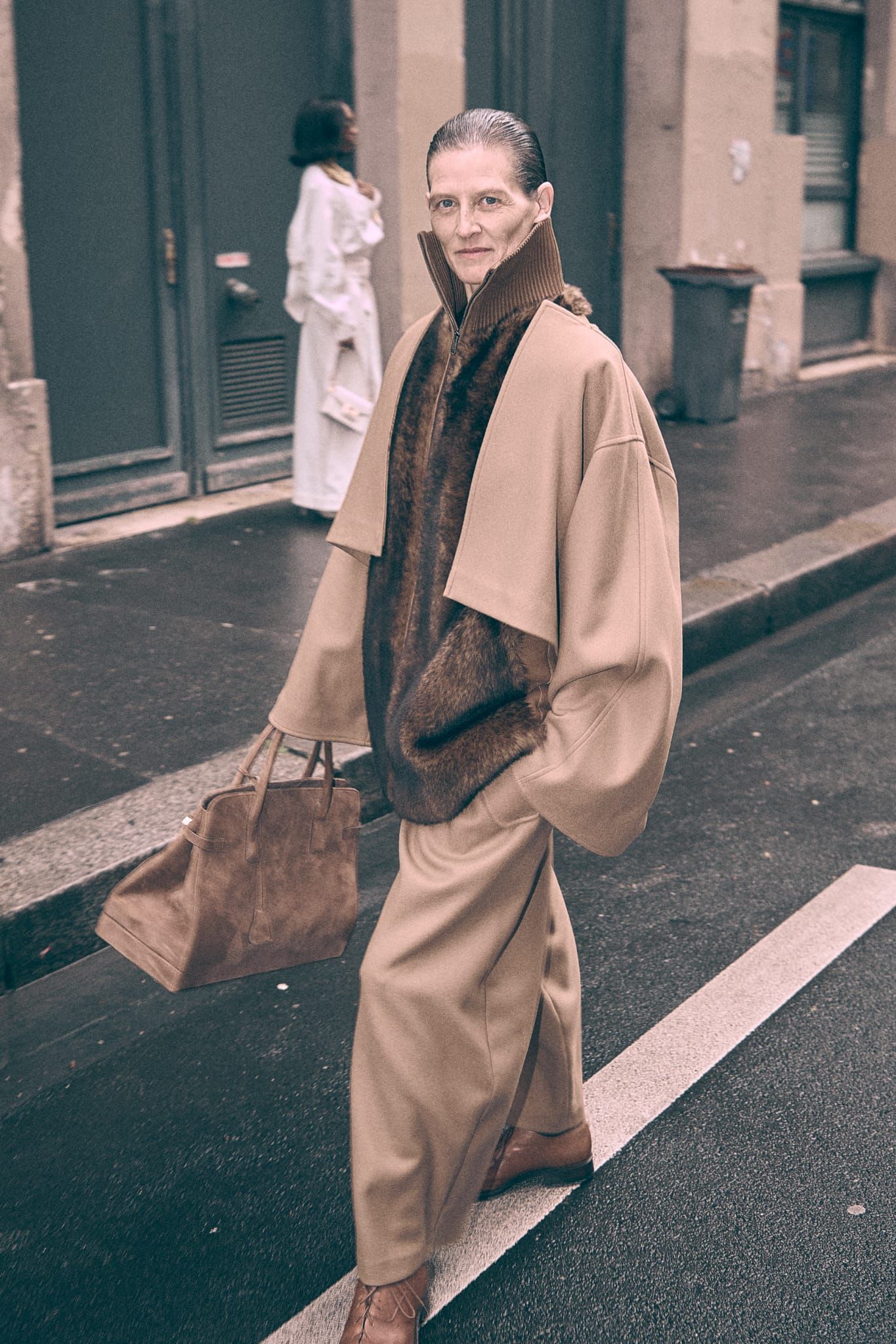 Una persona vestida con abrigo y pantalones color beige cruza una calle mojada, llevando una bolsa. Otra persona, con bata blanca, la sigue.